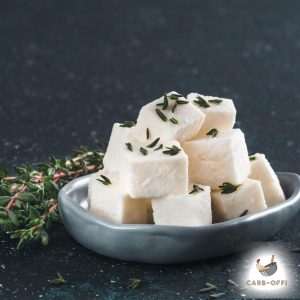 Small white cheese cubes in a dark grey oval plate with some oregano spread on top and some oregano leaves next to the plate