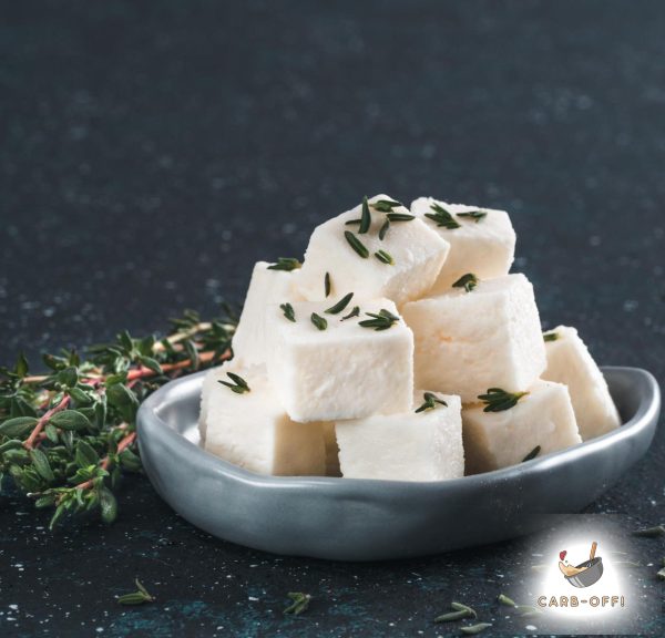 Small white cheese cubes in a dark grey oval plate with some oregano spread on top and some oregano leaves next to the plate