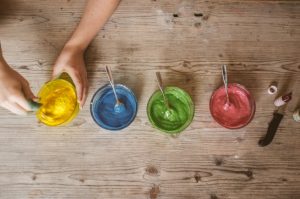 Four glass jars with different colours of natural food colouring with a spoon in each one of them. First colour from left is yellow, then blue, then green, then red. Woman's hand holding the side of the bowl with the yellow colouring