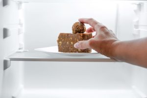 Black man's hand reaching out to a fridge shelf to grab a piece of chocolate cake with a chocolate truffle on top from a white plate