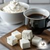 Black cup of black coffee on a wooden board behind five off white, square pieces of energy bombs and a white bowl filled up with whipped cream in the background