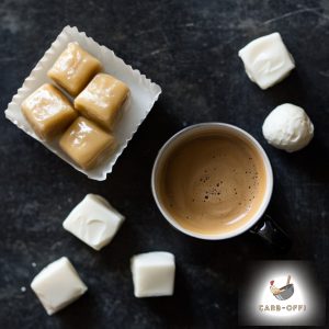 Photo taken from above of a cup of hot frothy coffee, next to a square white plate with 4 pieces of buttery caramel in it and 5 white square & round fat bombs randomly placed next to it