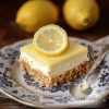 Square piece of a lemon curd cheesecake - biscuit layer at the bottom, cream layer in the middle, topped with lemon curd and a slice of lemon on a vintage plate with a silver teaspoon