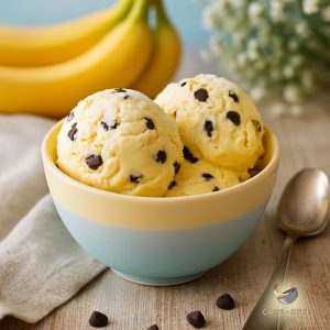 Two scoops of banana choc chip ice cream in a bowl with pastel colours on a rustic, wooden table with a few choc chips around the bowl and a bunch of bananas in the background