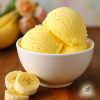 Three scoops of banana ice cream in a white bowl with a few banana slices next to it on a wooden board and a bunch of bananas showing up blurred in the background