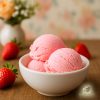 Three scoops of strawberry ice cream in a white bowl on a wooden table next to fresh strawberries with a vase of pastel coloured flowers in the background
