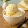 Three scoops of plain vanilla ice cream in a white bowl on a wooden board next to two long , black vanilla beans and a vase with flowers and a pastel colour towel in the background