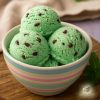 Three scoops of vivid green coloured ice cream with choc chips, in a pastel coloured bowl on a wooden table