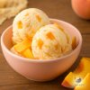 Two big scoops of off white ice cream with peach tidbits in a pastel pink bowl on a wooden table next to peach slices
