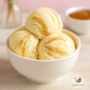 Three scoops of salted caramel ice cream with caramel syrup streaks on a white wooden table with a bowl of liquid caramel in the background