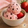 Two scoops of light pink strawberry choc chip ice cream in an off white bowl on wooden table with a couple of fresh strawberries in the background