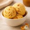 Three scoops of pale orange coloured ice cream with chunks of butterscotch fudge in a white bowl on a wooden table and a couple butterscotch pieces next to it